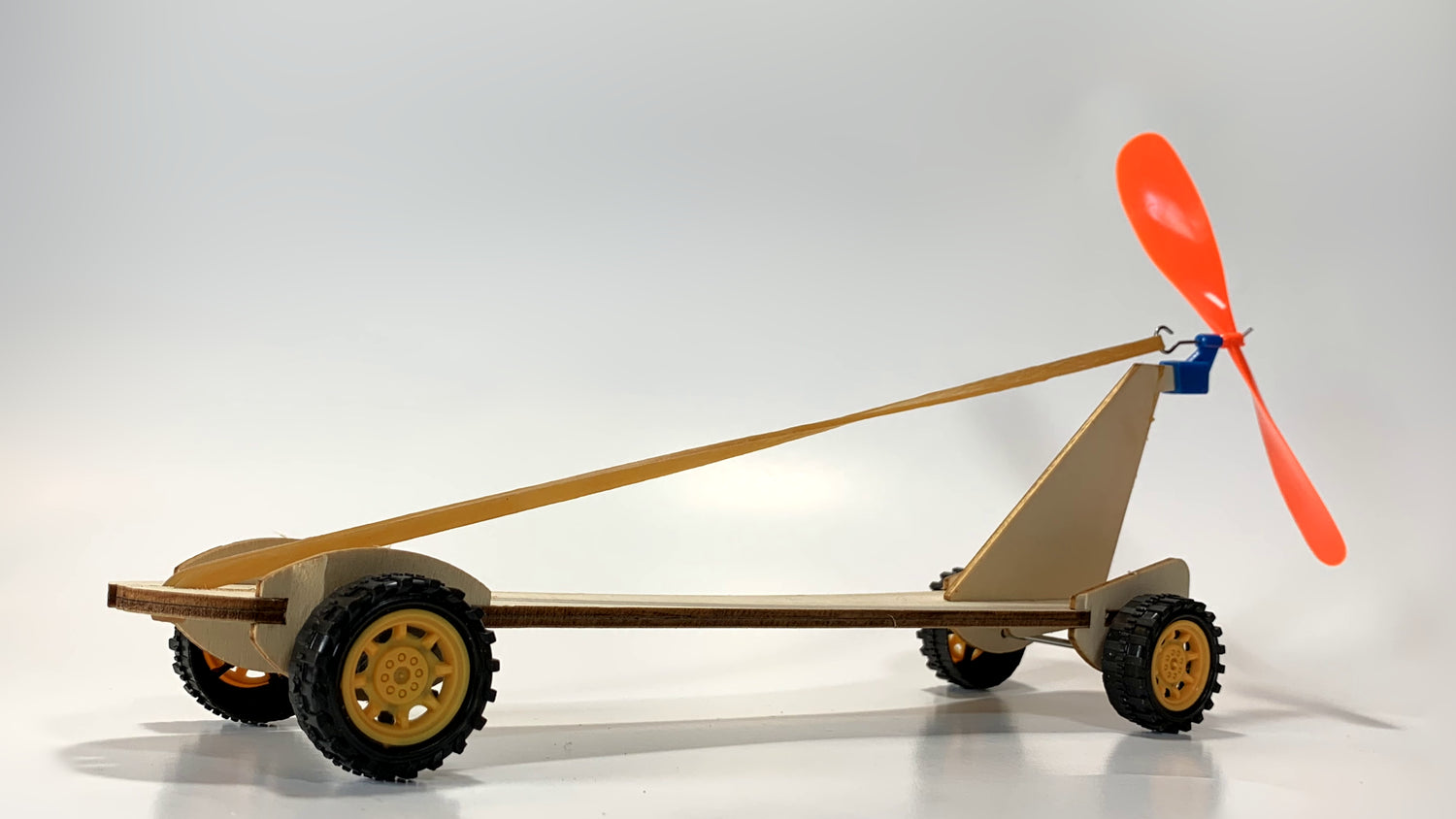 Alternate side view of the Rubber Band Race Car STEM Kit featuring a wooden frame, yellow wheels, elastic rubber band, and bright orange propeller