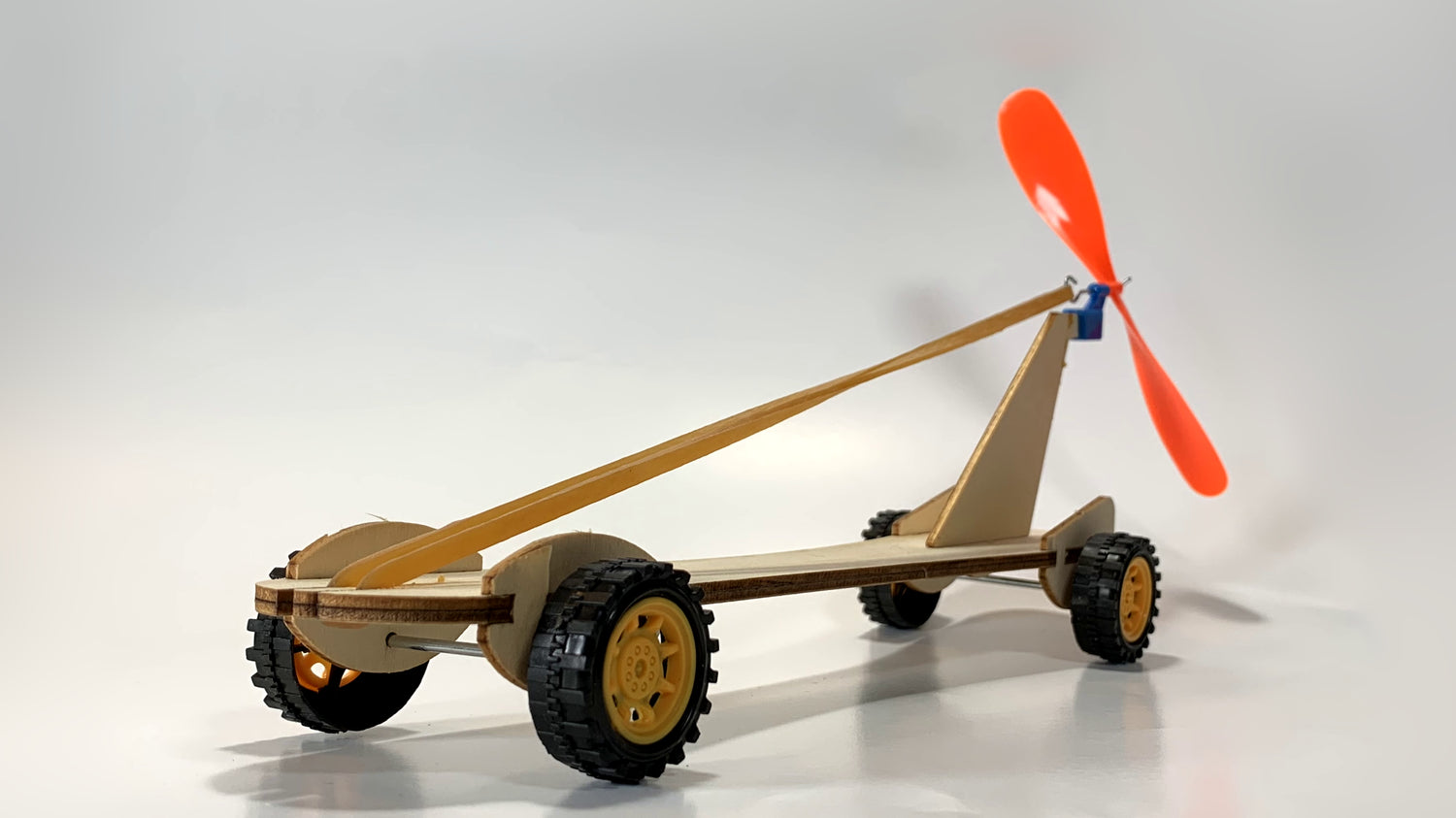 Rear angle view of the Rubber Band Race Car STEM Kit featuring a wooden frame, yellow wheels, stretched rubber band, and bright orange propeller