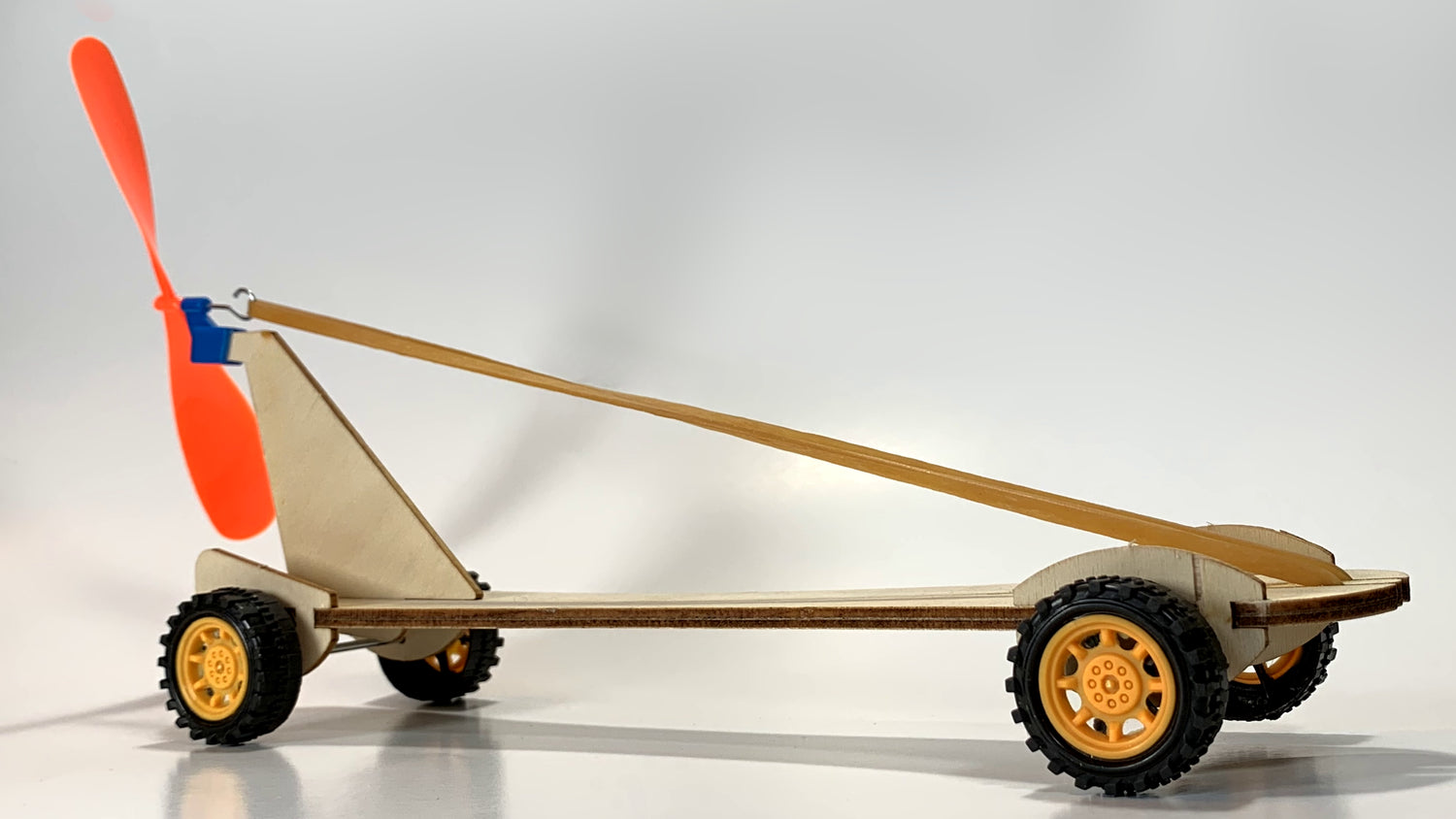 Side view of the Rubber Band Race Car STEM Kit showing a wooden frame, yellow wheels, stretched rubber band, and bright orange propeller